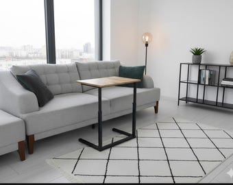 Puede incluir: Un salón moderno con un sofá gris claro y cojines verdes oscuros y negros. Una pequeña mesa de madera ajustable con patas negras se encuentra frente al sofá. Una alfombra geométrica en blanco y negro y una estantería de metal negro completan la decoración.