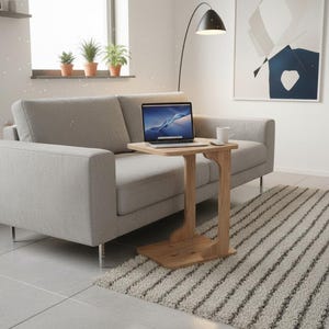 May include: A light gray sofa with a wooden side table holding a laptop and a mug. The table has a unique, curved design. A rug with black and white stripes is on the floor. The room has a modern, minimalist aesthetic.