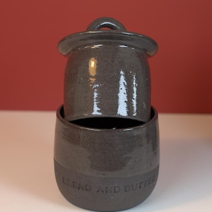 Peut inclure: Un beurrier en céramique gris foncé avec un couvercle et une base. La base porte les mots "BREAD AND BUTTER" en relief. Le plat est sur une surface blanche avec un fond rouge.