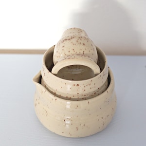 Può includere: Un set di tre ciotole da portata in ceramica maculata color crema. Le ciotole sono impilate, con un piccolo recipiente a forma di brocca all'interno. I pezzi di ceramica hanno un aspetto rustico e fatto a mano, con macchie marroni visibili.