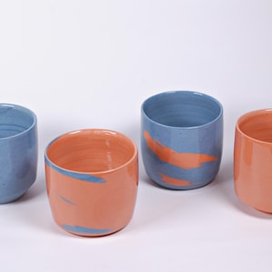 Op de afbeelding: Vier keramische bekers in blauwe en oranje tinten. Twee zijn blauw met oranje accenten, en twee zijn oranje met blauwe details. De bekers hebben een gladde, glanzende afwerking en een eenvoudig, modern ontwerp.