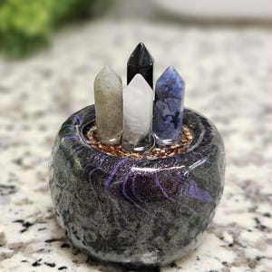 May include: A small, round, decorative pot with a marbled black and purple design, holding four crystal points. The crystals are in shades of gray, white, black, and blue. The pot sits on a speckled surface.