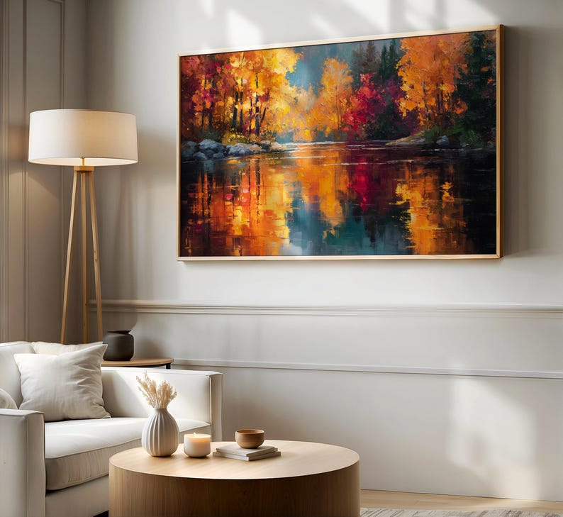 May include: A framed landscape painting depicts a vibrant autumn scene. The artwork showcases a river reflecting the colorful trees in shades of red, orange, and yellow. The painting is displayed above a white sofa and round wooden coffee table.