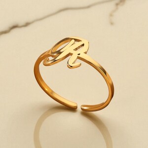 Könnte beinhalten: Ein goldfarbener, verstellbarer Ring mit dem Buchstaben "R" in Schreibschrift. Der Ring ist unten offen und spiegelt sich auf der darunter liegenden Oberfläche. Der Ring ist ein Schmuckstück.