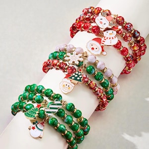 Op de afbeelding: Verschillende armbanden met kerstthema bedels. De armbanden hebben rode, groene en witte kralen, met bedels waaronder een sneeuwpop, de kerstman en een kerstboom. De armbanden worden op een wit oppervlak weergegeven.