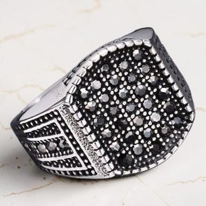 Op de afbeelding: Een zilverkleurige metalen ring met een brede, rechthoekige kop die dicht bezet is met talloze kleine, gefacetteerde zwarte stenen. De ringband heeft een textuurrijke, geoxideerde afwerking met geometrische patronen en zwarte accenten.