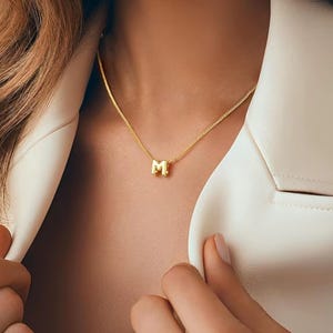 Könnte beinhalten: Eine goldfarbene Halskette mit einem Anhänger in Form des Buchstabens "M". Die Halskette wird zu einem weißen Blazer getragen. Die Kette ist filigran und der Anhänger ein Blockbuchstabe. Das Model hat lange, wellige, hellbraune Haare.