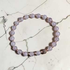May include: A beaded bracelet featuring smooth, pale lavender round beads alternating with small metallic rose gold spacer beads. A single spherical accent bead encrusted with tiny black stones is positioned near the bottom of the circular strand.