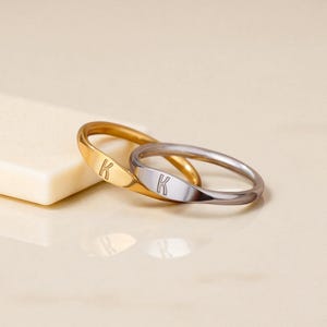 Könnte beinhalten: Zwei Ringe, einer goldfarben und einer silberfarben, jeweils mit dem Buchstaben "K" auf einer rechteckigen Platte graviert. Die Ringe sind ineinander verschlungen und liegen auf einer hellen Oberfläche. Der goldfarbene Ring hat eine polierte Oberfläche.