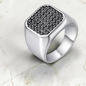 Può includere: Un anello a sigillo color argento con una superficie quadrata incastonata con una griglia di piccole pietre rotonde nere. La fascia è liscia, lucida e presenta una struttura ampia e solida che si restringe leggermente verso la base.