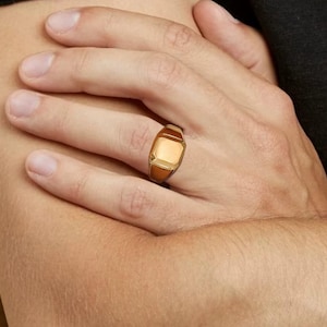 Könnte beinhalten: Ein goldfarbener Siegelring mit rechteckiger Oberfläche. Der Ring hat eine polierte Oberfläche und wird am Finger getragen. Das Design des Rings weist eine geometrische Form und eine warme Farbpalette auf.
