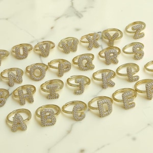 Könnte beinhalten: Eine Sammlung von goldfarbenen Metallringen, von denen jeder einen einzelnen Buchstaben des Alphabets trägt, der mit kleinen, klaren, funkelnden Steinen besetzt ist. Die Ringe sind verstreut angeordnet und zeigen das gesamte Alphabet von A bis Z.