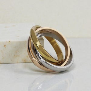 Puede incluir: Un anillo multicolor con tres bandas entrelazadas en oro, oro rosa y plata. Las bandas son lisas y pulidas, creando un diseño entrelazado y sin costuras. El anillo se muestra sobre una superficie blanca.