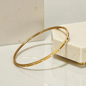 May include: A gold-colored bangle bracelet with small, clear gemstones evenly spaced around the band. The bracelet is circular and sits on a white surface, with a white and beige background.