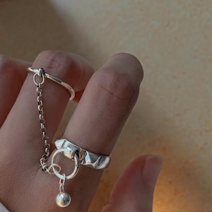 Puede incluir: Un conjunto de anillos de plata con dos anillos conectados por una delicada cadena. Uno es una banda simple, mientras que el otro tiene un diseño geométrico y una pequeña esfera colgante. La joya está diseñada para usarse en dos dedos.