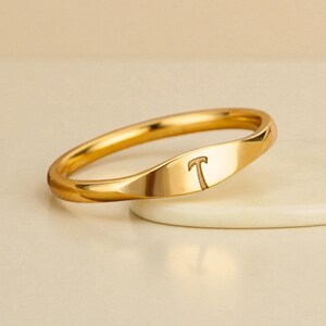 Könnte beinhalten: Ein goldfarbener Ring mit poliertem Finish. Der Ring hat einen flachen, rechteckigen Abschnitt mit dem eingravierten Buchstaben "T". Der Ring ist etwa 0,6 cm breit und hat ein glattes, geschwungenes Design.