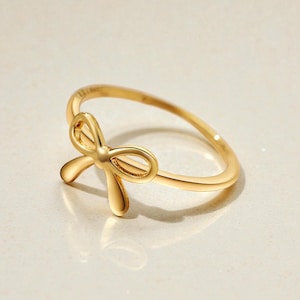 Puede incluir: Un anillo dorado con un diseño de lazo. El anillo tiene una banda delgada y un lazo en relieve. El lazo está centrado en el anillo y tiene un acabado liso y pulido. El anillo se muestra sobre una superficie de color claro.