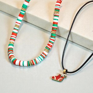 Op de afbeelding: Twee kettingen. Eén is een kralenketting met rode, witte, groene en gouden kralen. De andere is een zwarte koordketting met een rood-witte kerstsokhanger met gouden accenten. De kettingen liggen op een lichtgrijze ondergrond.