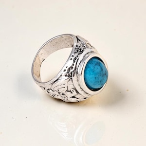 Op de afbeelding: Een zilverkleurige metalen ring met een ovaalvormige, felblauwe steen met donkere aders in het midden. De ringband vertoont een ingewikkeld, verhoogd reliëfpatroon met gestructureerde details over het gehele buitenoppervlak.
