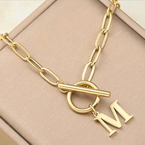 Op de afbeelding: Goudkleurige ketting met een tuimelsluiting en een hanger met de letter "M". De ketting is gemaakt van ovale schakels. De ketting wordt gepresenteerd op een beige oppervlak.