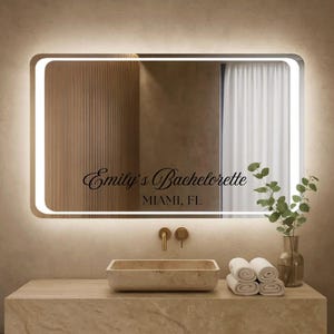 May include: A rectangular, illuminated mirror with rounded corners is mounted above a stone countertop. The mirror has the text "Emily's Bachelorette, Miami, FL" etched on its surface. A stone sink, gold fixtures, rolled white towels, and a vase with greenery complete the bathroom setting.