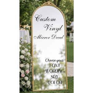 May include: An arched floor mirror with a gold frame featuring black vinyl lettering that reads Custom Vinyl Mirror Decal, followed by Choose your FONT WORDING SIZE COLOR. The mirror reflects an outdoor setting with green trees and white flowers.