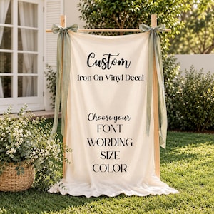 Op de afbeelding: Een crèmekleurige stoffen banner hangend aan een houten frame met saliegroene linten aan de bovenhoeken. De banner bevat zwarte tekst: Custom Iron On Vinyl Decal, Choose your FONT, WORDING, SIZE, COLOR.