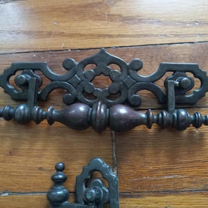 May include: Ornate, dark bronze cabinet hardware. The handles have a decorative, scrolling design with a central floral motif. The pulls are a dark brown color. The hardware is set against a wooden surface.