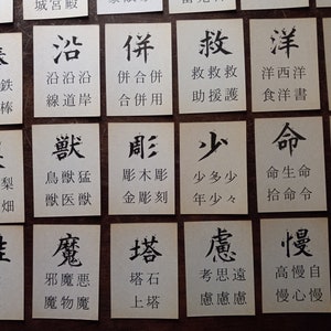 May include: A collection of small, square cards with black Japanese calligraphy on a wooden surface. Each card features a different character, with additional text below. The cards are arranged in a grid.