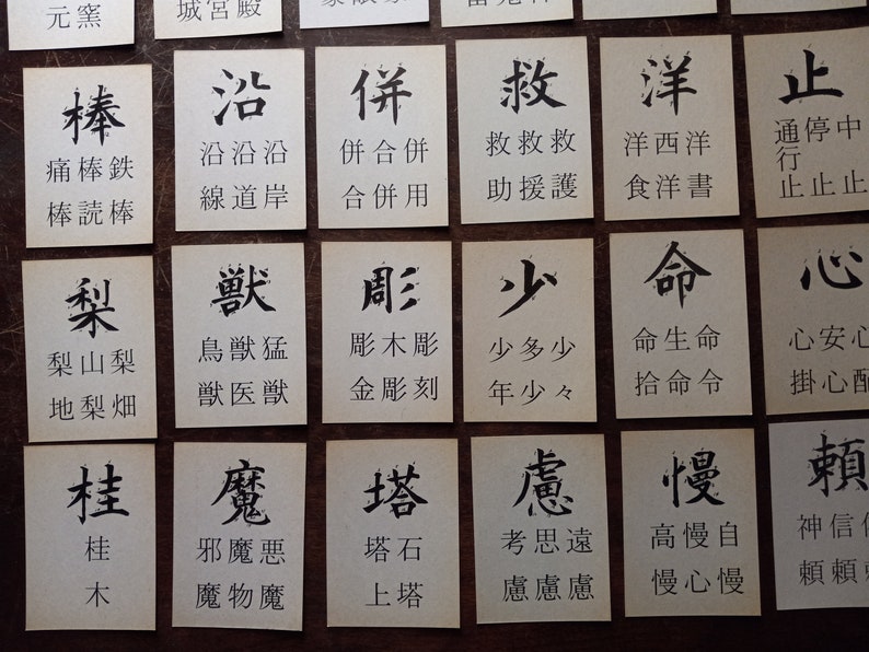 May include: A collection of small, square cards with black Japanese calligraphy on a wooden surface. Each card features a different character, with additional text below. The cards are arranged in a grid.