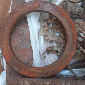 May include: A circular metal ring with a heavily oxidized, rust-colored surface. The object features a thick, flat profile and a weathered texture, showing signs of corrosion and age across its entire circumference and outer edge.