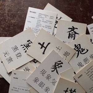 May include: A collection of vintage flashcards with Japanese characters and English definitions, scattered on a dark wooden surface. The cards feature black ink calligraphy and dictionary entries, creating a scholarly aesthetic.
