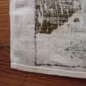 May include: Close-up of a white linen tea towel with a brown printed design. The towel has a hemmed edge.