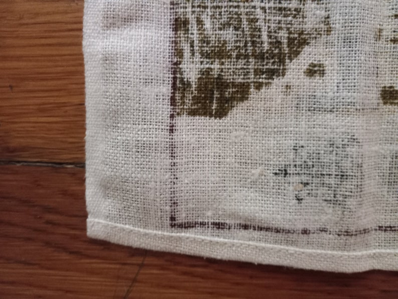 May include: Close-up of a white linen tea towel with a brown printed design. The towel has a hemmed edge.