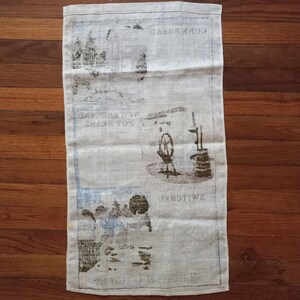 May include: A white linen tea towel with a brown and black printed design. The design features a windmill, a well, and the text "Swiss" and "Corn Mill".