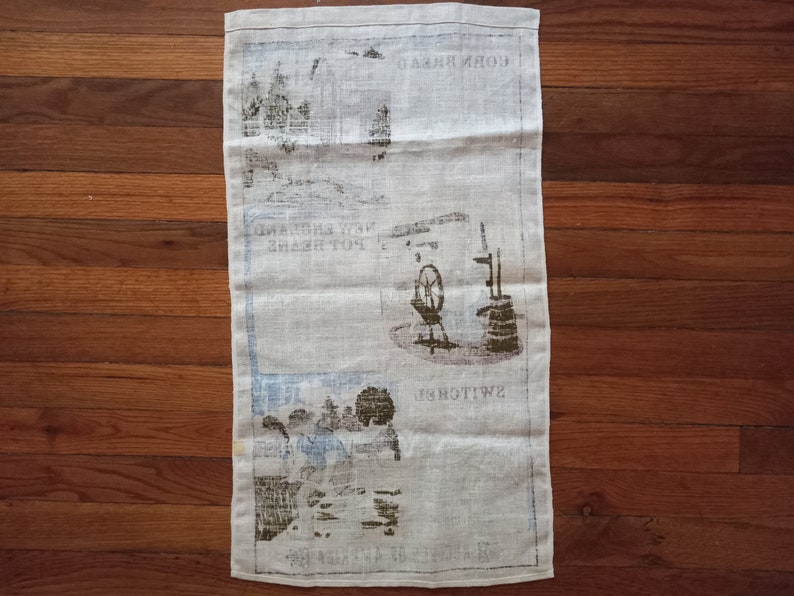 May include: A white linen tea towel with a brown and black printed design. The design features a windmill, a well, and the text "Swiss" and "Corn Mill".
