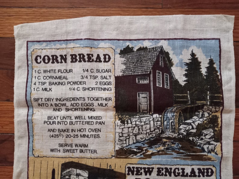 May include: A vintage kitchen tea towel with a recipe for cornbread printed on it. The towel features a rustic illustration of a New England farmhouse with a water wheel. The recipe includes ingredients like white flour, cornmeal, sugar, salt, baking powder, eggs, milk, and shortening. The text reads "CORN BREAD" and "NEW ENGLAND".
