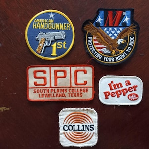 Pode incluir: Uma coleção de patches bordados com vários designs. Um patch apresenta uma arma de fogo e o texto "AMERICAN HANDGUNNER 1st". Outro mostra uma águia com o texto "PROTECTING YOUR RIGHT TO RIDE". Outros patches incluem "SPC SOUTH PLAINS COLLEGE LEVELLAND, TEXAS", "I'm a Pepper" e "COLLINS".