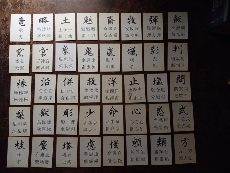 May include: A collection of small, rectangular cards with black Japanese characters printed on a white background. The cards are arranged on a dark surface, likely for a game or educational purpose.