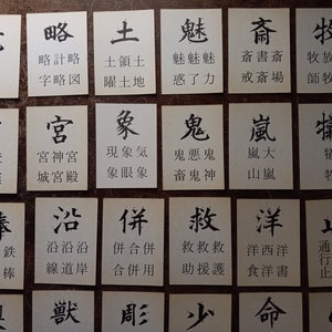 May include: A collection of small, white cards with black Japanese characters and their phonetic transcriptions. The cards are arranged in a grid pattern on a brown surface. The characters are written in a traditional calligraphic style.