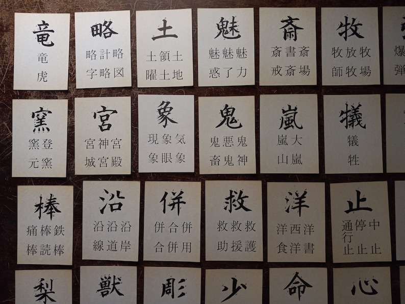 May include: A collection of small, white cards with black Japanese characters and their phonetic transcriptions. The cards are arranged in a grid pattern on a brown surface. The characters are written in a traditional calligraphic style.