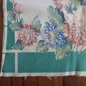 May include: A rectangular fabric textile featuring a white background with a large floral print of pink, red, and blue chrysanthemums and green leaves. The design is framed by a solid teal green border with white grid lines along the edges.