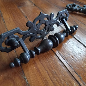 May include: Two ornate, dark metal cabinet handles with decorative scrollwork and a turned, bulbous handle design. The handles are set against a warm, wooden surface, showcasing their antique aesthetic.