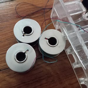May include: Three silver sewing machine bobbins with thread in various colors, including red, green, and black. The bobbins are on a wooden surface, with a clear plastic bobbin holder in the background. The bobbins are used for sewing.