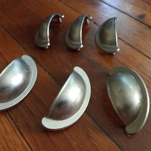 May include: A set of six silver-toned metal cup pulls for drawers or cabinets. Each pull features a smooth, semi-circular design with a curved handle and flat mounting flanges on either side, displaying a metallic finish with subtle variations.