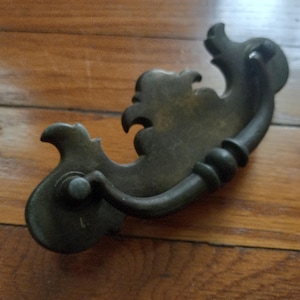 May include: Antique bronze-toned drawer pull with ornate, scroll-like detailing. The handle features a curved design with decorative elements and a circular base for mounting. A vintage hardware piece.