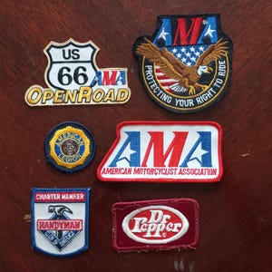 Pode incluir: Coleção de patches bordados com vários designs e logotipos. Inclui um patch da Rota 66 dos EUA, uma águia com uma bandeira americana e patches para a American Motorcyclist Association e Dr. Pepper.