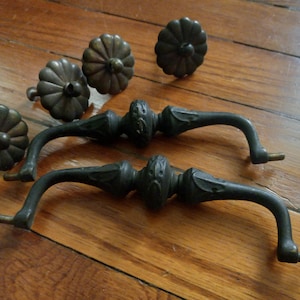 May include: Antique bronze-toned cabinet hardware set, including four decorative knobs and two ornate pulls. The knobs have a flower-like design, and the pulls feature detailed carvings. The hardware is displayed on a wooden surface.