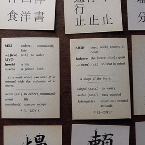 May include: Close-up of several small, rectangular cards with Japanese text and English translations. The cards are arranged on a wooden surface. The cards feature words and their meanings, such as "MEI" and "SHIN".