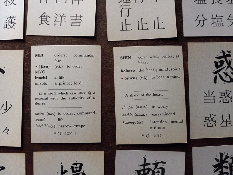 May include: Close-up of several small, rectangular cards with Japanese text and English translations. The cards are arranged on a wooden surface. The cards feature words and their meanings, such as "MEI" and "SHIN".